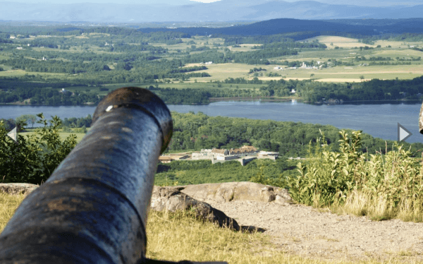 A historic cannon points toward a river and the distant Fort Ticonderoga, surrounded by lush green trees and fields, with mountains visible in the background under a clear sky.